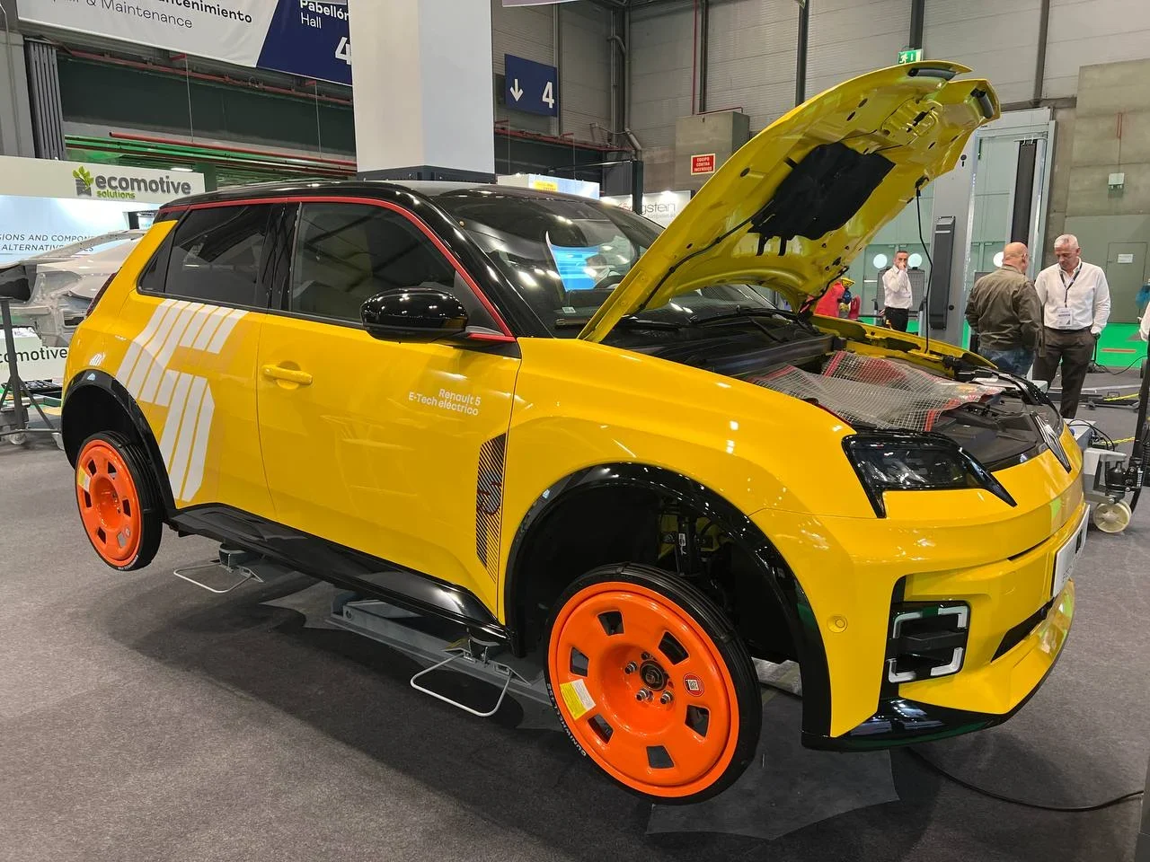 Un automóvil amarillo y negro con ruedas naranjas se exhibe con el capó abierto en una exposición.