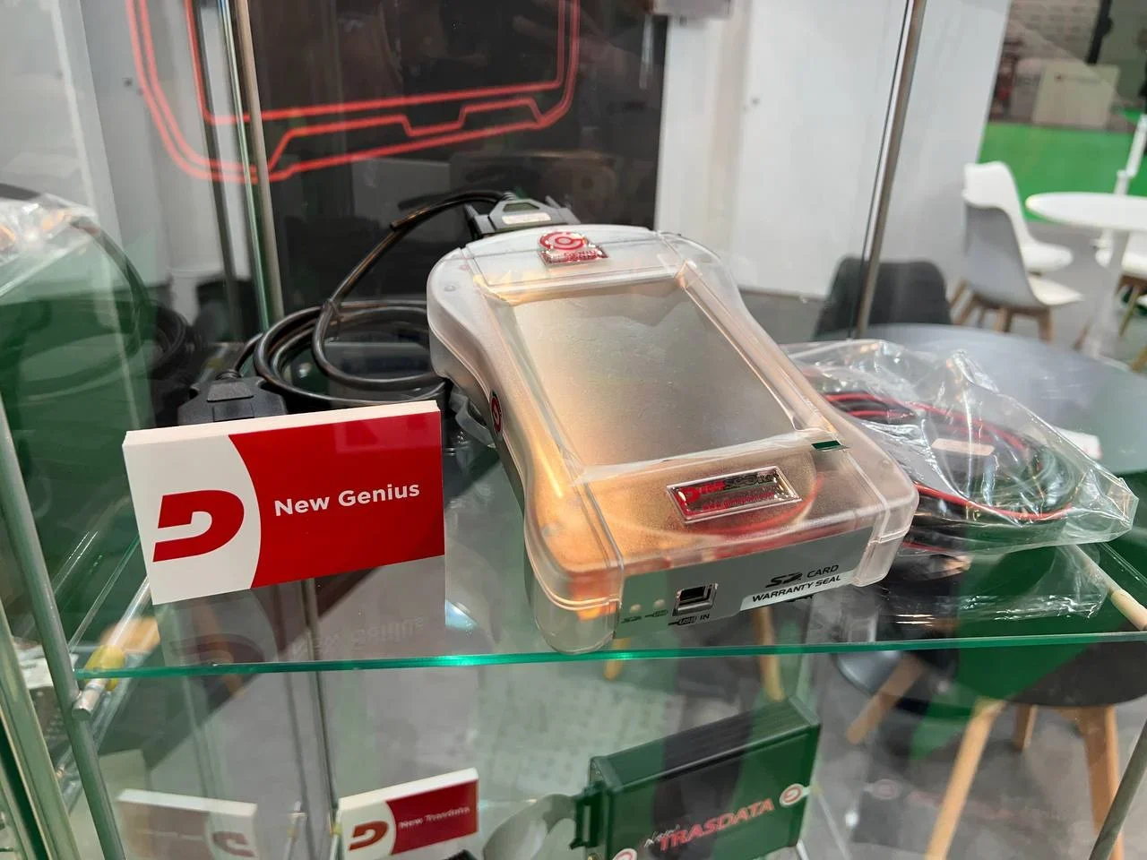 Dispositivo electrónico de plástico transparente con la etiqueta "New Genius" exhibido en un estante de vidrio con cables al lado.