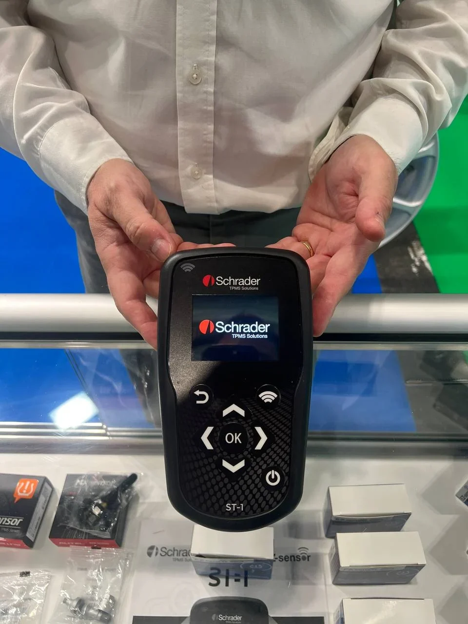 Persona sosteniendo una herramienta de diagnóstico TPMS de Schrader encima de una vitrina de vidrio con accesorios y embalaje.