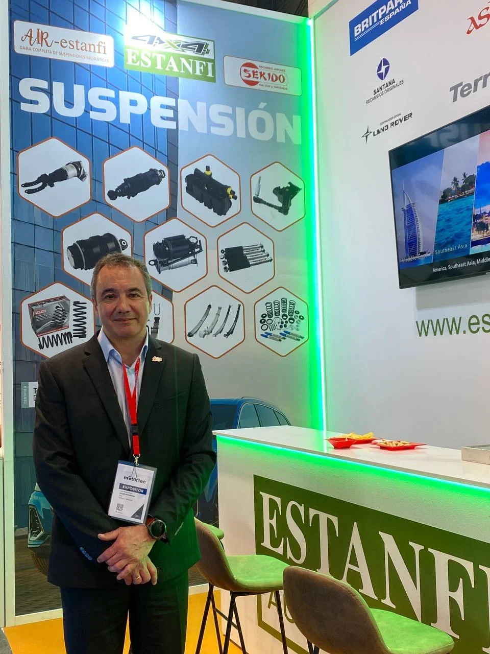 Un hombre con traje se encuentra en un stand de suspensiones automotrices en una feria comercial, sonriendo a la cámara.