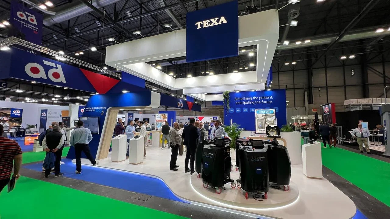 La gente camina e interactúa en un concurrido stand de feria de tecnología con la marca TEXA y pantallas de visualización.