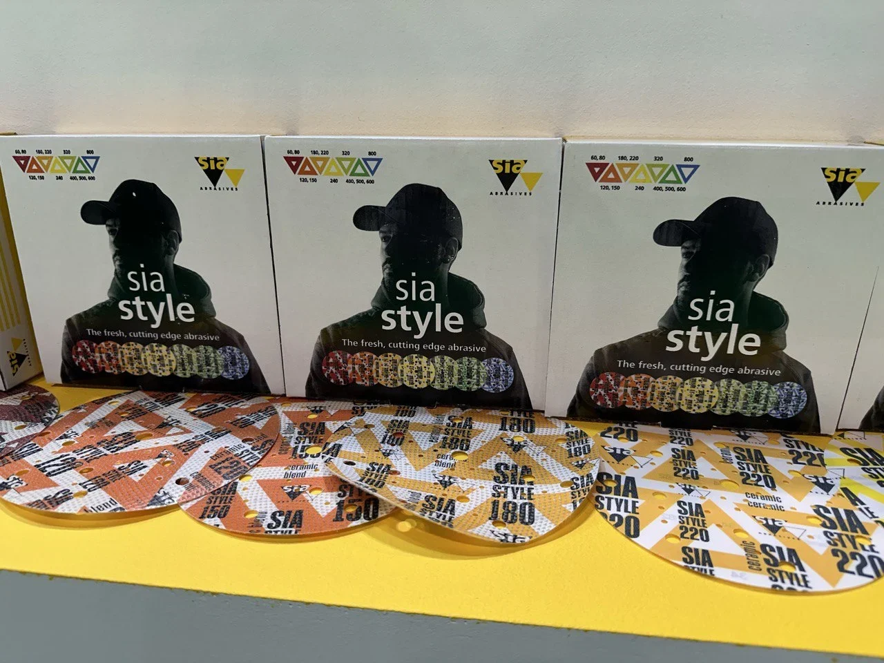 Tres cajas de discos abrasivos "estilo sia" exhibidas con varios discos dispuestos frente a ellas en un estante amarillo.