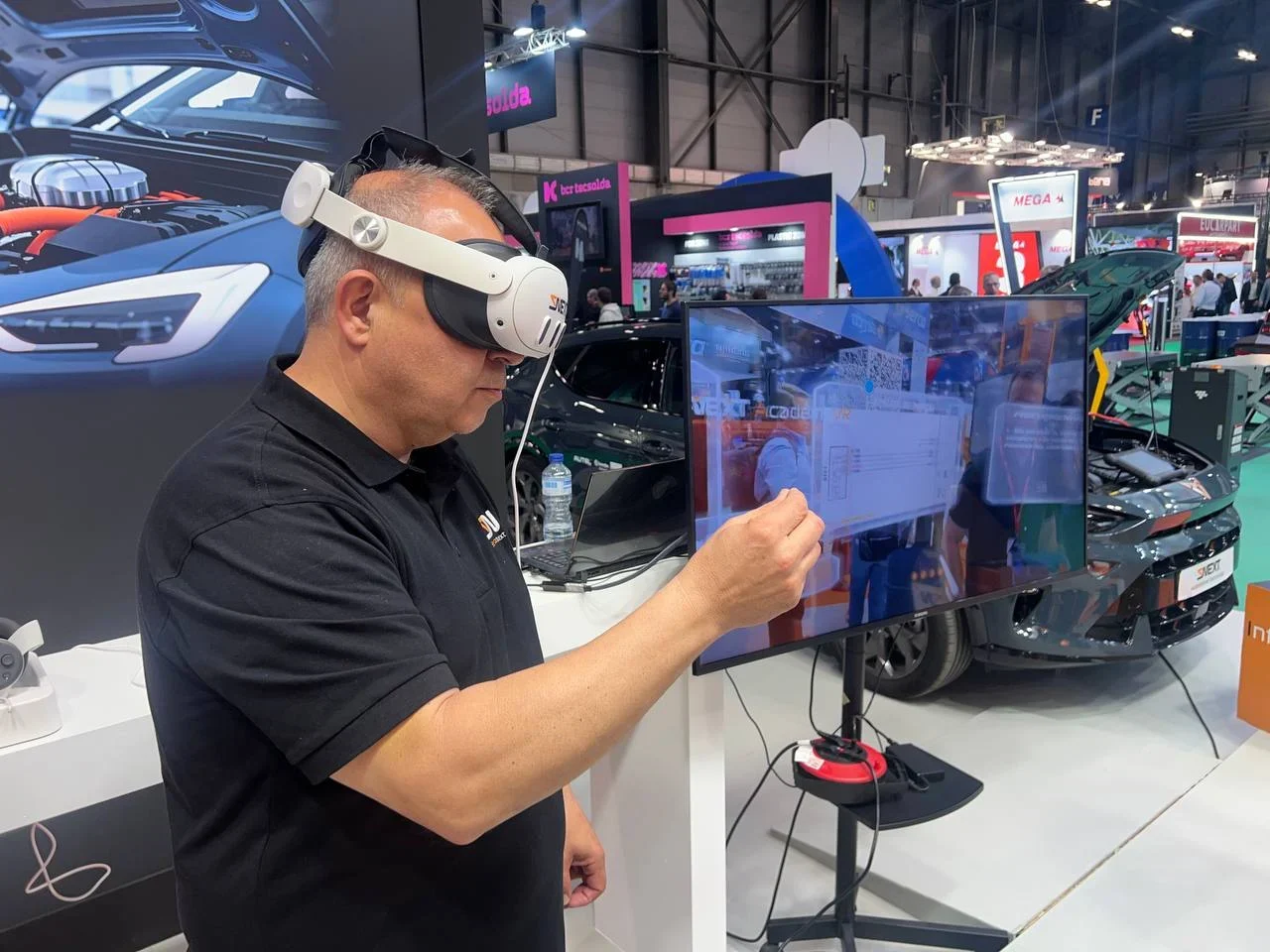 Un hombre que usa un casco de realidad virtual interactúa con una pantalla virtual en un stand de una exposición tecnológica con automóviles en el fondo.