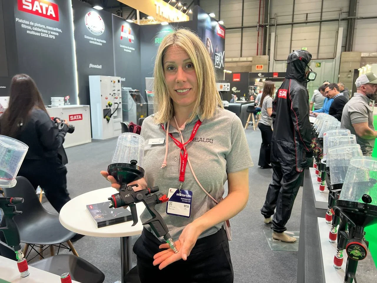 Una mujer en un stand de una feria comercial muestra una pistola pulverizadora de pintura, sonriendo a la cámara.