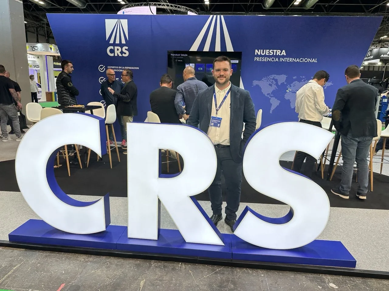 Un hombre se encuentra frente a unas grandes letras "CRS" en un stand de un evento empresarial mientras hay gente conversando detrás de él.