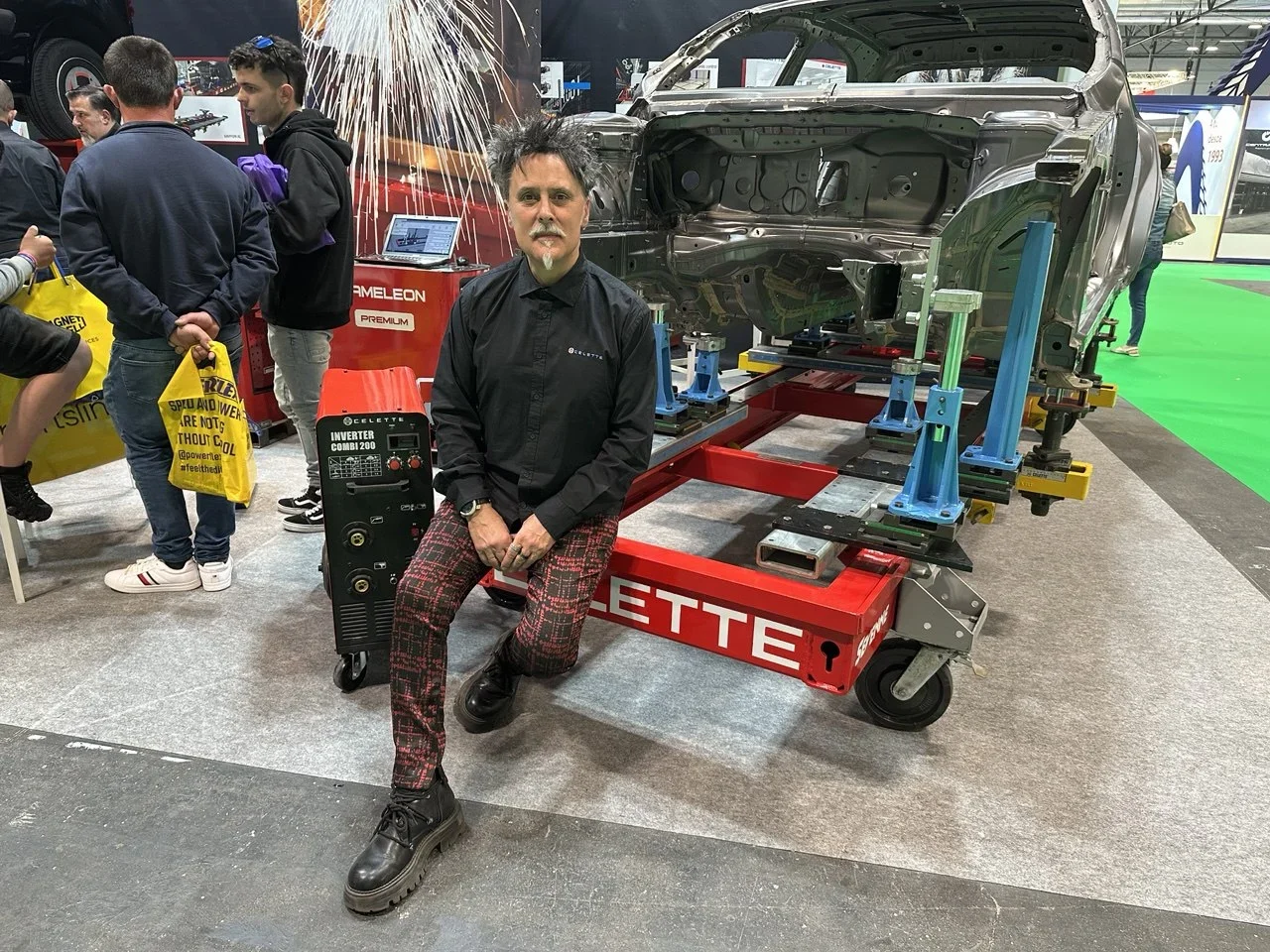 Un hombre se sienta en una máquina de soldar al lado de un chasis de automóvil desmontado en una exposición automotriz en interiores.