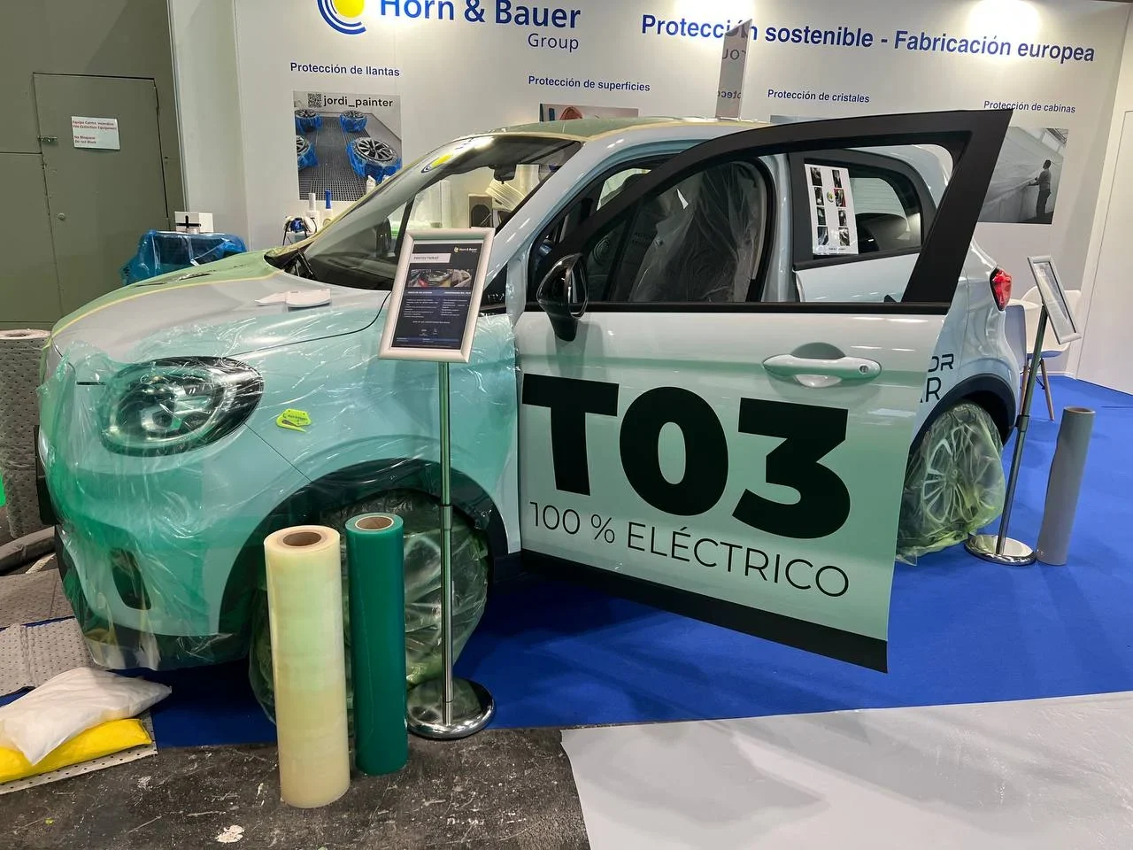 Un automóvil eléctrico verde etiquetado "T03 100% Eléctrico" se exhibe en un stand de exposición con envoltorios protectores.