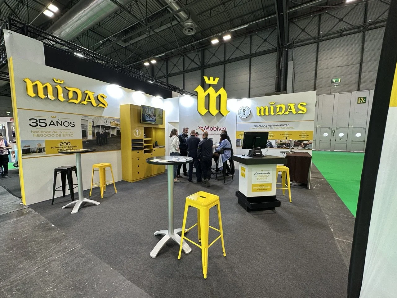 Stand de exposición de Midas con detalles en amarillo, taburetes, pantalla de televisión y gente conversando en el interior.