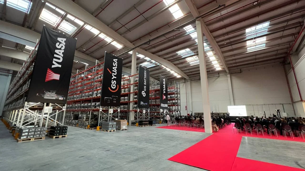 Un almacén con estantes, pancartas, alfombra roja y filas de sillas preparadas para un evento o presentación.
