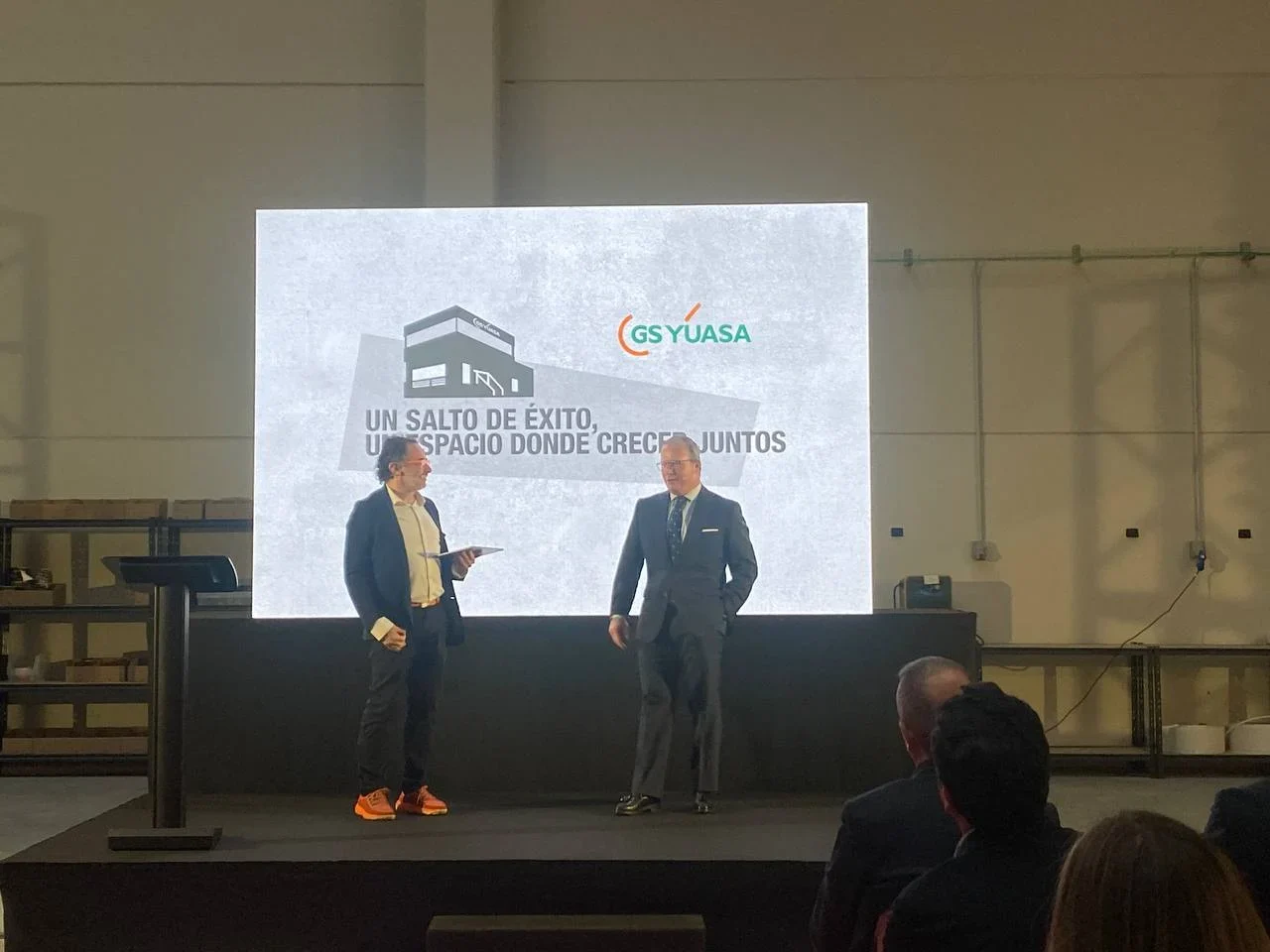 Dos hombres de traje se encuentran en el escenario frente a una pantalla con el logotipo de GS Yuasa y texto en español.