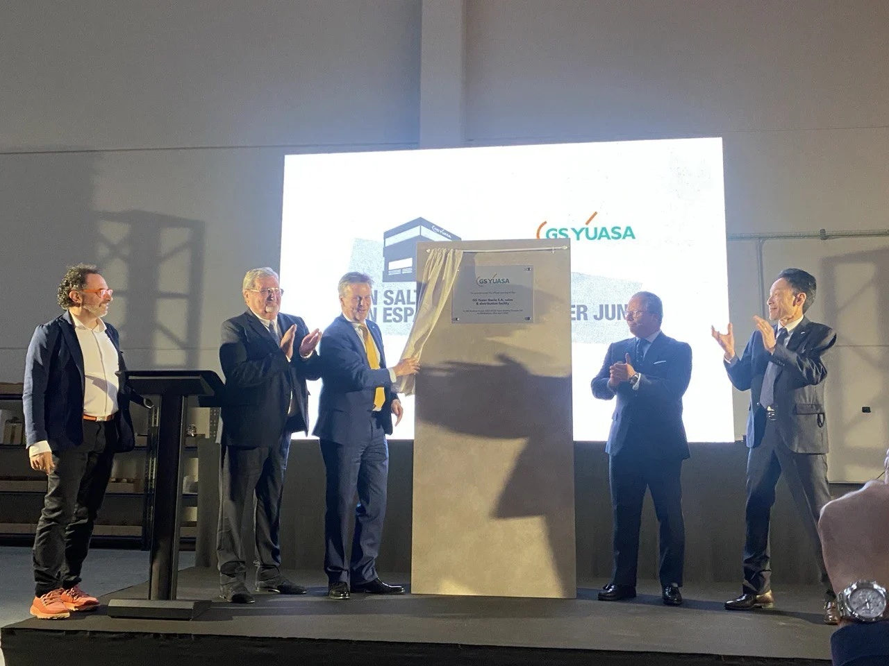 Cinco hombres de traje descubren una placa conmemorativa en una ceremonia en el interior, aplaudiendo y sonriendo.