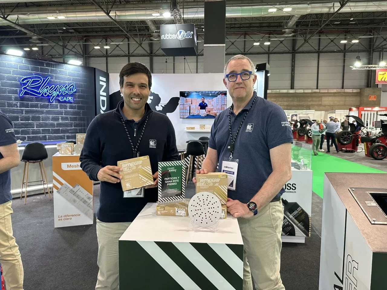 Dos hombres con camisas a juego se encuentran junto a una exhibición de productos en una feria comercial, sosteniendo discos de lijado.