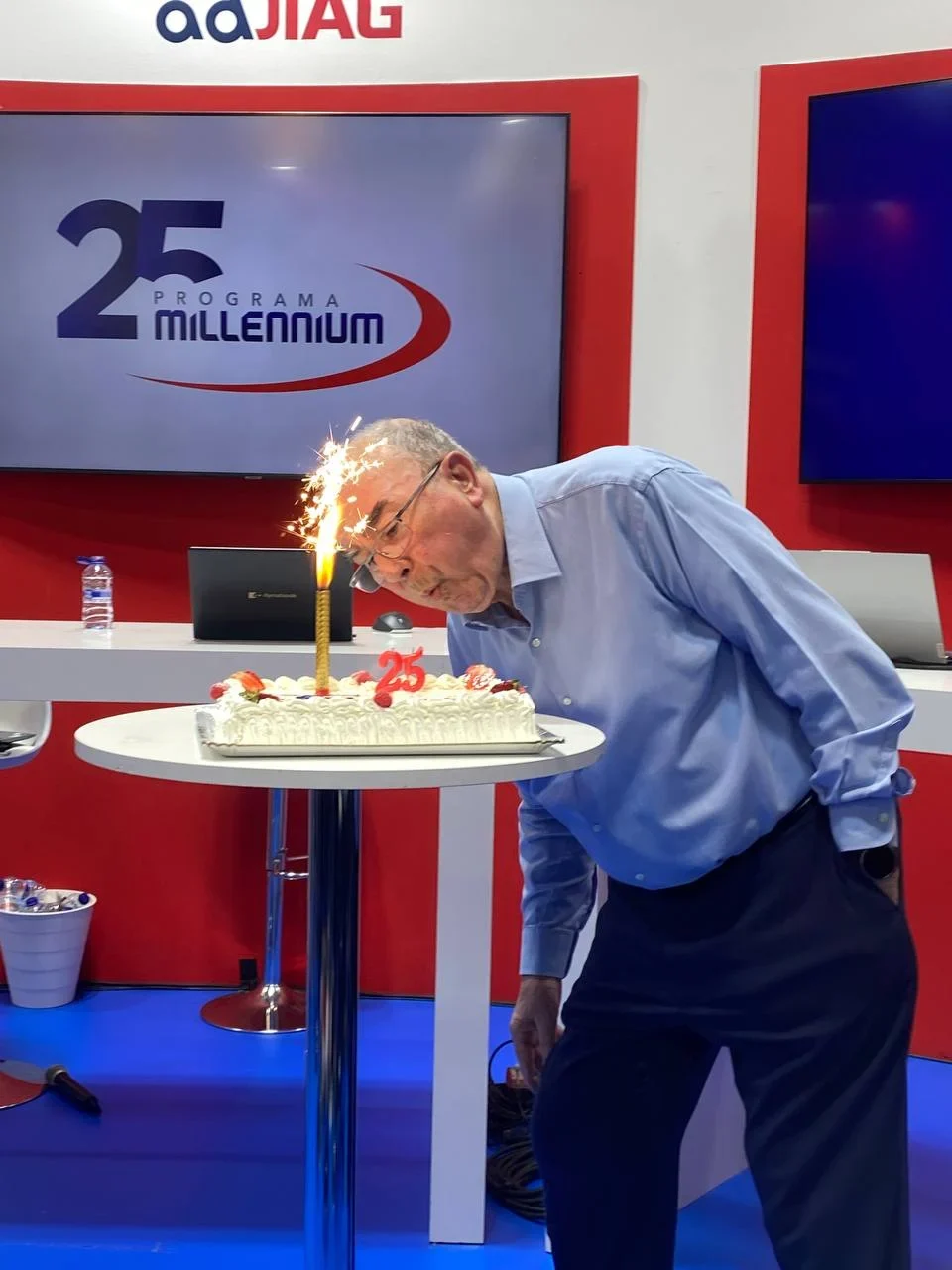 Un hombre mayor se inclina para soplar una vela brillante en un pastel que dice "25" en un evento de celebración.