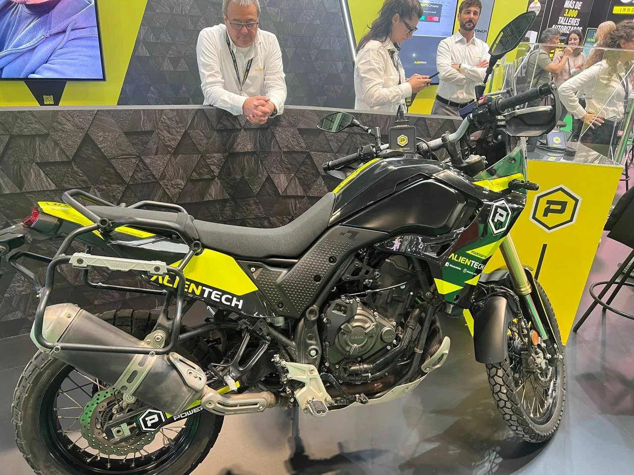 Una motocicleta de aventura amarilla y negra en exhibición en el interior, con gente de pie observando en el fondo.