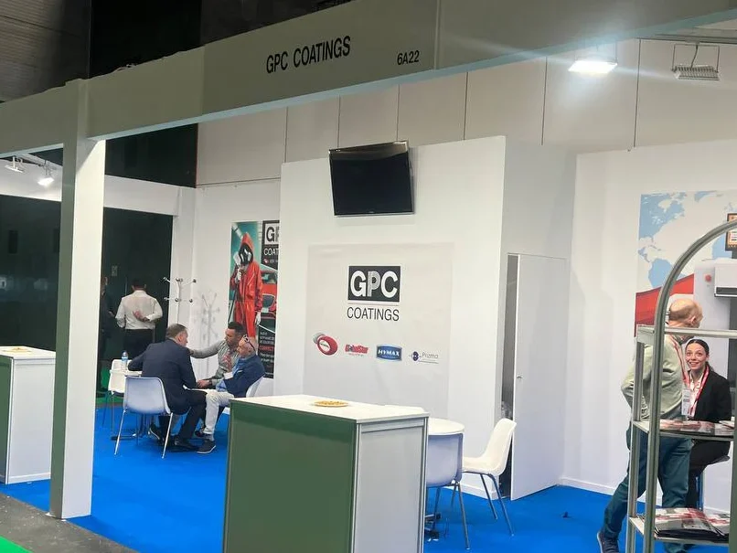 Tres hombres se sientan y conversan en un stand de feria comercial de GPC Coatings, con paredes blancas y una alfombra azul brillante.