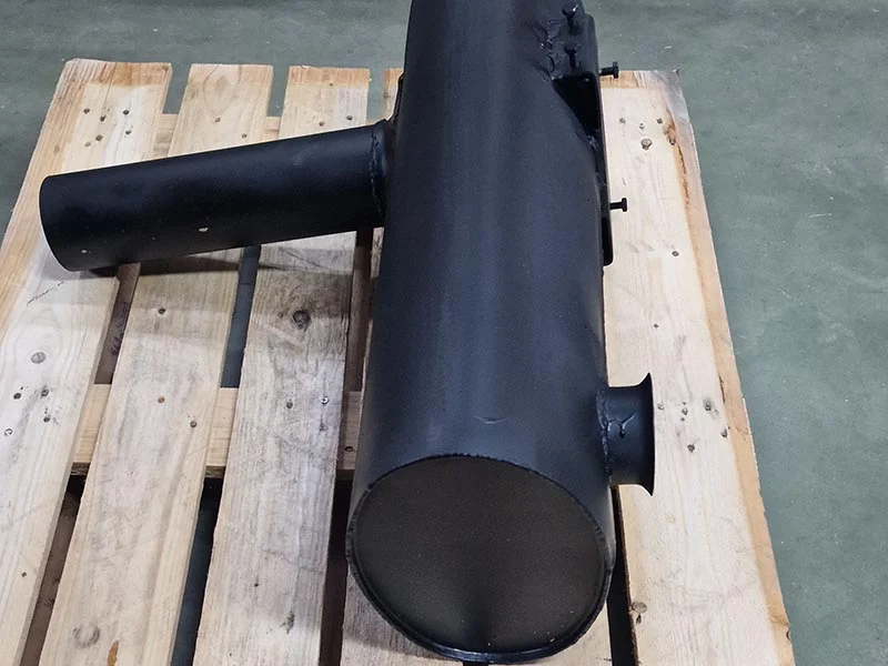 Un silenciador de metal negro, fabricado para sistemas de escape de vehículos, se coloca sobre una paleta de madera sobre un piso de concreto.