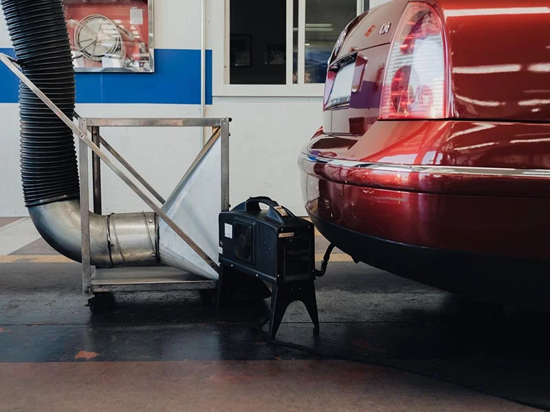 Un dispositivo prueba las emisiones del tubo de escape de un automóvil rojo en un taller mecánico cerrado: menos humo, más planeta.