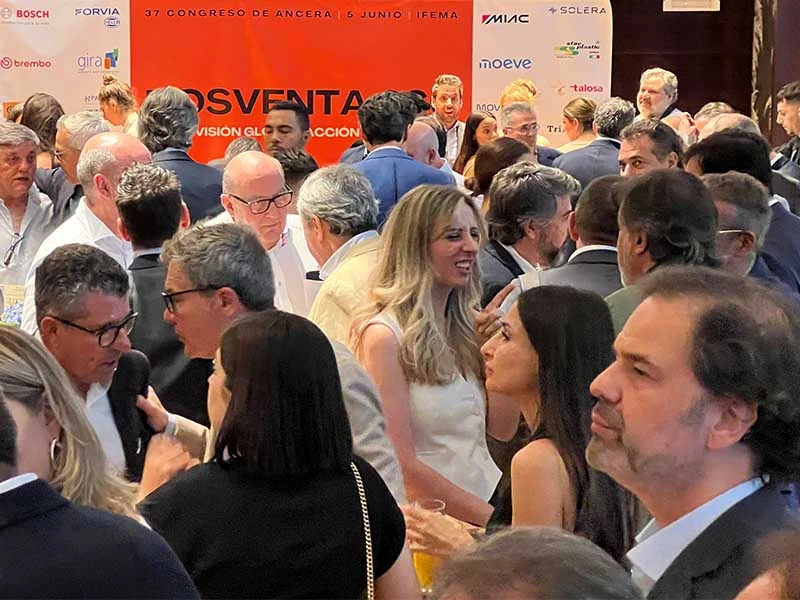 Multitud de personas haciendo networking en un evento profesional, con pancartas y logotipos visibles en el fondo.