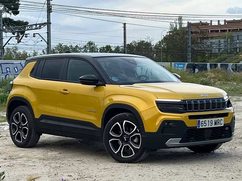 Un todoterreno Jeep amarillo estacionado al aire libre sobre una superficie de tierra, con edificios y líneas eléctricas al fondo.
