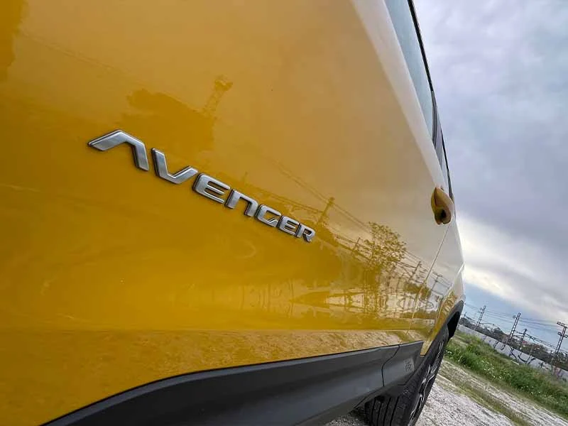 Primer plano de un automóvil amarillo con la insignia "AVENGER" en el costado, estacionado al aire libre en un día nublado.
