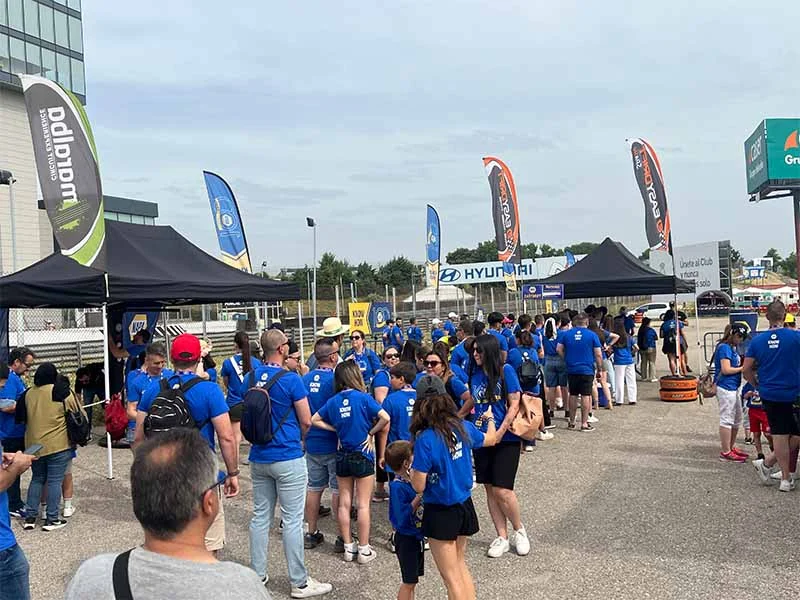 Un gran grupo de personas con camisas azules se reúne al aire libre cerca de tiendas de campaña y banderas en el evento NAPA Jarama, creando un animado ambiente de fiesta.