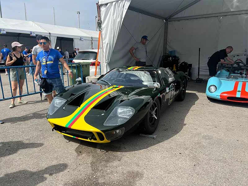 Un coche de carreras clásico con franjas amarillas, verdes y rojas está aparcado cerca de una carpa en una exposición de coches en el Jarama, lo que se suma al ambiente festivo de fiesta.
