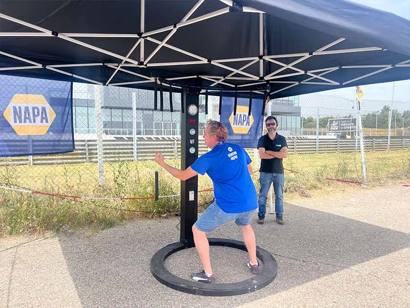 En una animada fiesta en el Jarama, una persona utiliza una máquina de reflejos de boxeo al aire libre bajo una carpa, mientras otra sonríe y observa.