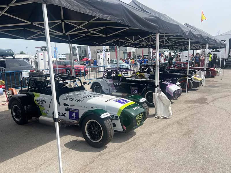 Cuatro coches de carreras, incluido un colorido Fiesta, aparcados bajo marquesinas en un evento de automovilismo en el Jarama, con gente y otros coches al fondo.