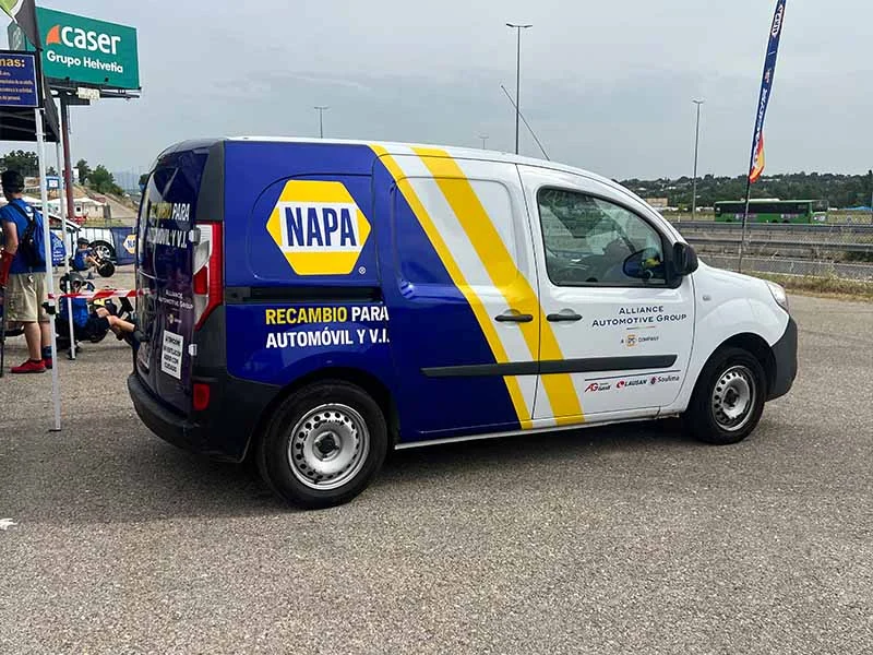 Una furgoneta de recambios NAPA con la marca azul y amarilla está estacionada en un estacionamiento cerca de una carretera y de gente, aportando un toque del espíritu festivo del Jarama a la escena.