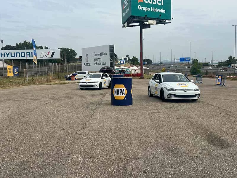 Dos coches blancos circulan a toda velocidad alrededor de un barril de NAPA en un gran estacionamiento abierto, evocando la emoción de una fiesta con carteles y espectadores de fondo.