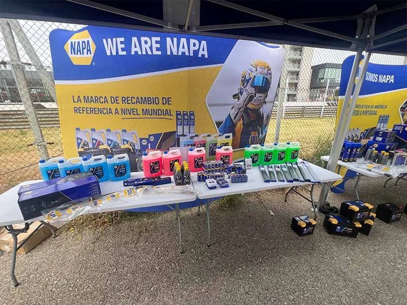 Una mesa de exhibición NAPA con productos automotrices y una pancarta brinda un ambiente festivo a un evento al aire libre, instalado junto a una cerca y una carretera.