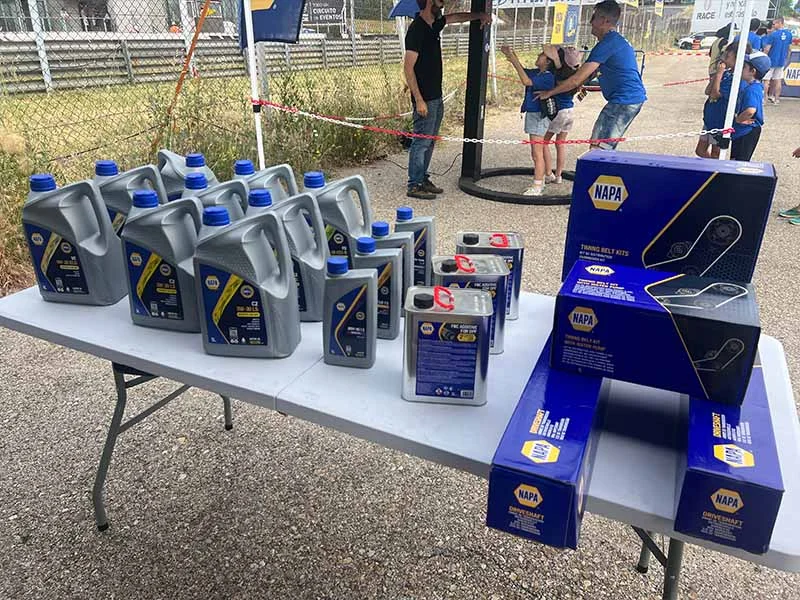 Mesa con contenedores y cajas de aceite de motor NAPA en un evento al aire libre, con gente con camisas azules disfrutando del ambiente de fiesta en el fondo.