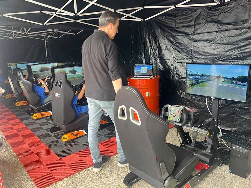 Varias personas disfrutan de simuladores de carreras bajo una carpa en el Jarama, como parte del animado ambiente de fiesta, con un hombre de pie cerca observando la acción.