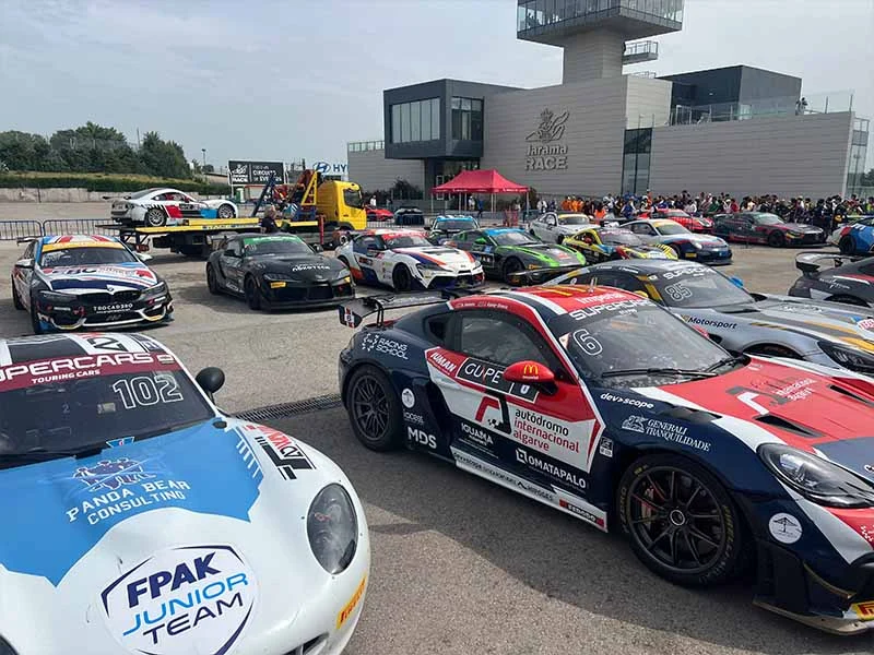 Un grupo de coches de carreras aparcados delante de un edificio moderno en el evento de deportes de motor del Jarama, creando un vibrante ambiente de fiesta.