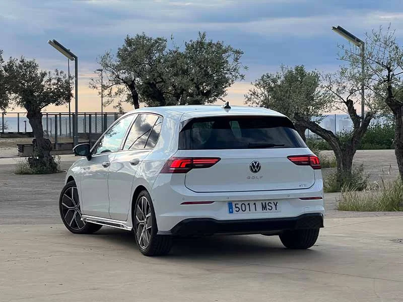 Un Volkswagen Golf R-Line 1.5 eTSI blanco estacionado al aire libre, cerca de árboles y bancos, visto desde atrás en un día nublado. Eterno referente en estilo e innovación.