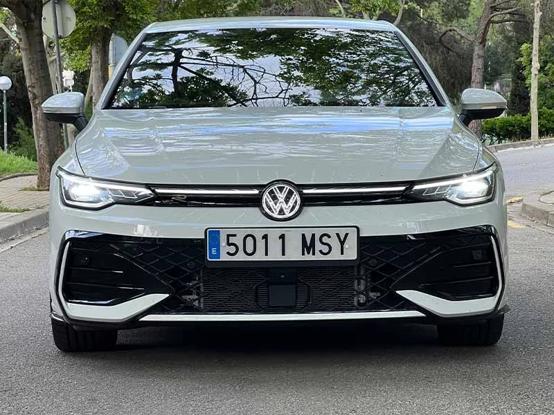 Vista frontal de un Volkswagen Golf R-Line 1.5 eTSI blanco estacionado en una calle, con árboles reflejados en el capó y el parabrisas: una eterna referencia de estilo e innovación.