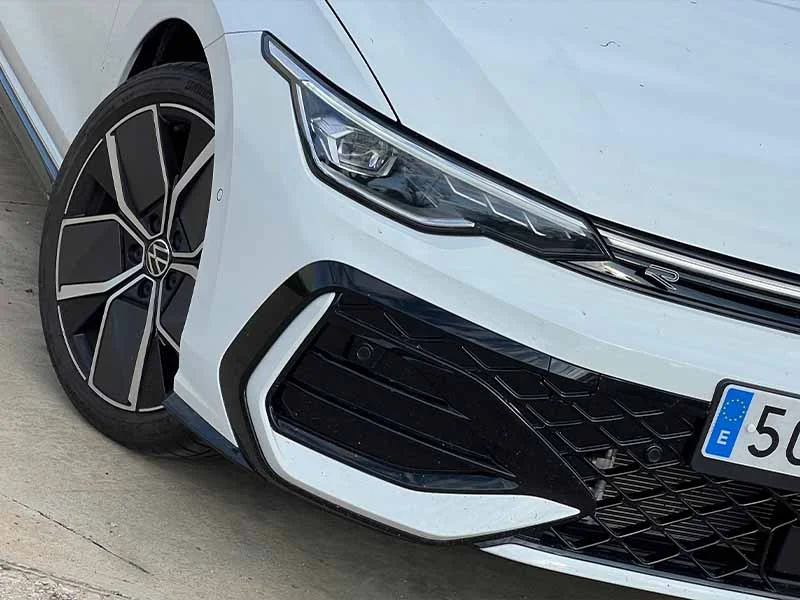 Primer plano de la parte delantera izquierda de un Golf R-Line blanco, con el faro, la parrilla y la llanta de aleación: un eterno referente impulsado por el avanzado motor 1.5 eTSI.