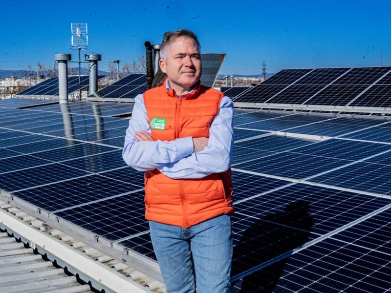 Un hombre con chaleco naranja se encuentra en una azotea con paneles solares, mostrando sostenibilidad bajo un cielo azul claro en RIC Madrid.