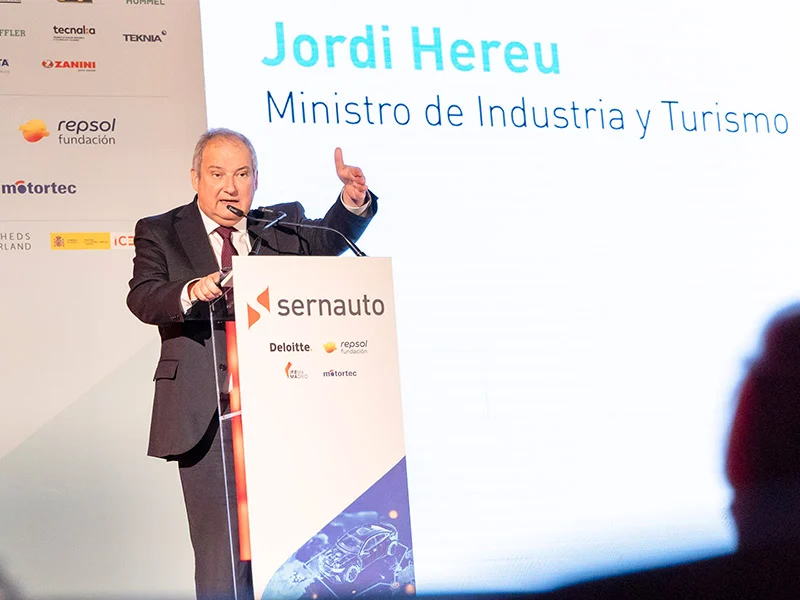 Un hombre trajeado habla en un podio durante un Encuentro, con una pantalla detrás en la que se lee "Jordi Hereu, Ministro de Industria y Turismo", destacando la colaboración entre SERNAUTO y ADN España.