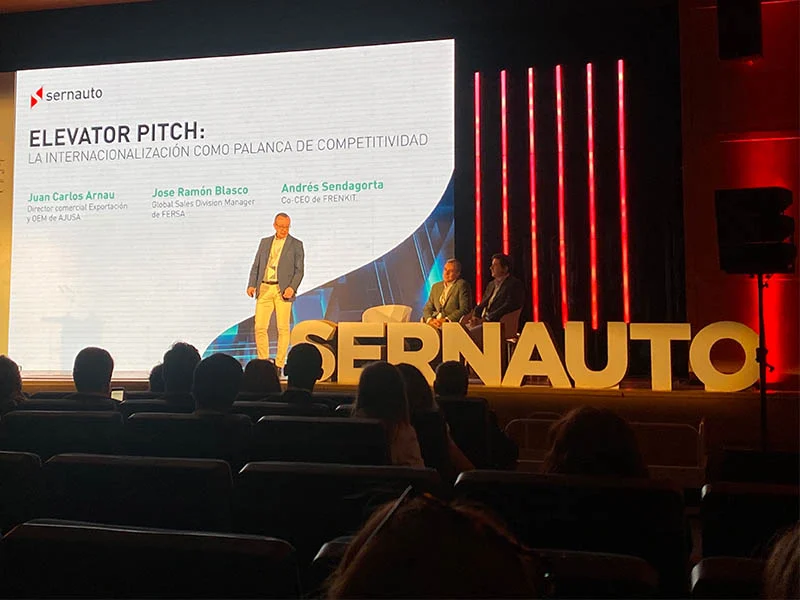 Tres hombres en el escenario de un encuentro empresarial con letras "SERNAUTO" y una presentación sobre internacionalización, destacando el espíritu ADN España.