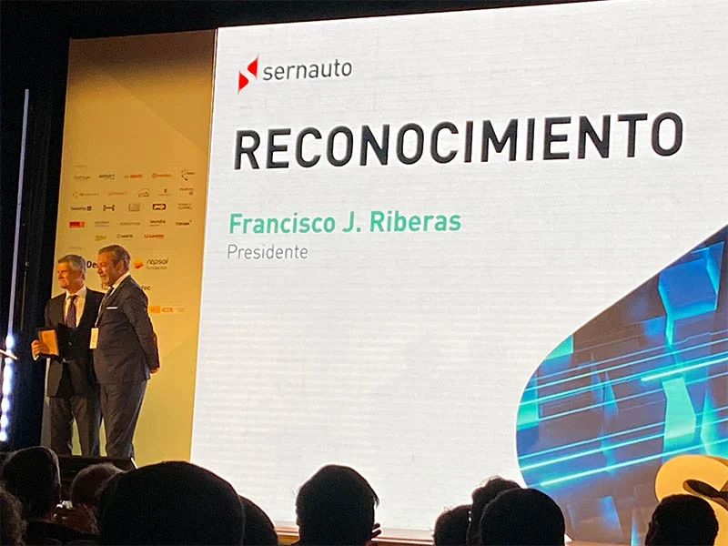 Dos hombres en el escenario de un acto de reconocimiento durante el Encuentro ADN España, con una gran pantalla que muestra "RECONOCIMIENTO", organizado por SERNAUTO.