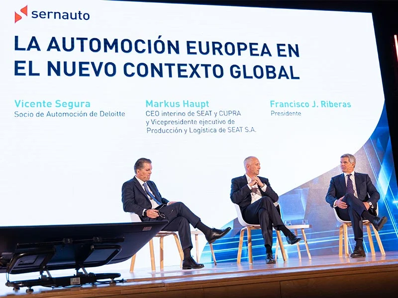 Tres hombres trajeados se sientan en el escenario para hablar de la automatización europea en una conferencia del Encuentro SERNAUTO, con una pantalla detrás.