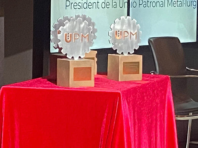 Sobre una mesa cubierta de terciopelo rojo, dos trofeos metálicos en forma de engranaje con la inscripción "UPM" celebran el espíritu de la Jornada del Metal 2025.