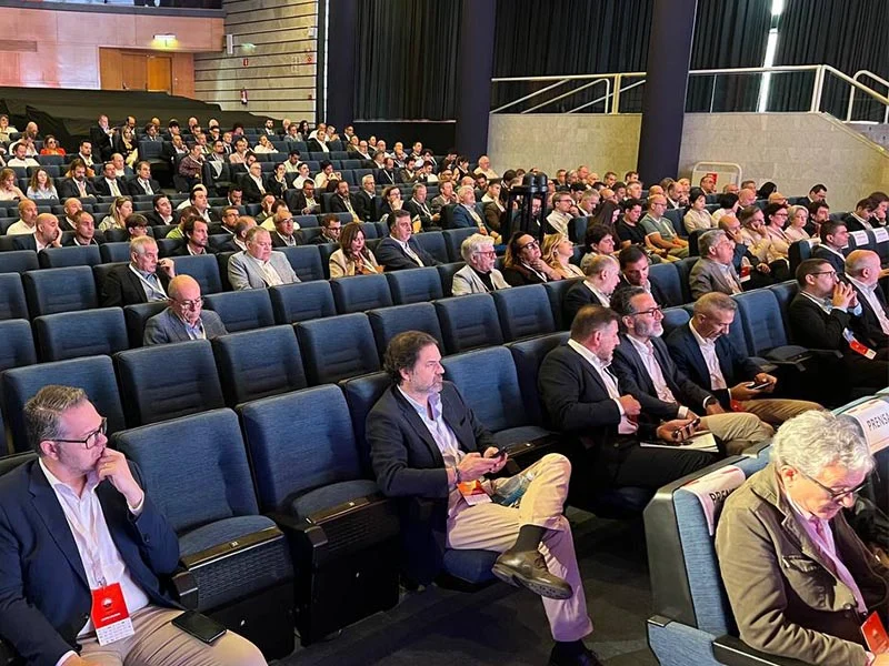 Un gran grupo de personas socializando en un evento empresarial con pancartas y logotipos en el fondo.