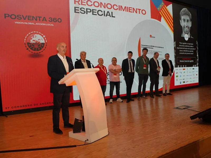 Un grupo de personas se encuentra en el escenario durante una ceremonia de premiación, con un hombre hablando en un podio y haciendo referencia al borrador automático durante su discurso.