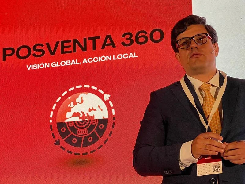 Hombre de traje presentando frente a una pantalla roja que decía "POSVENTA 360, VISIÓN GLOBAL, ACCIÓN LOCAL.