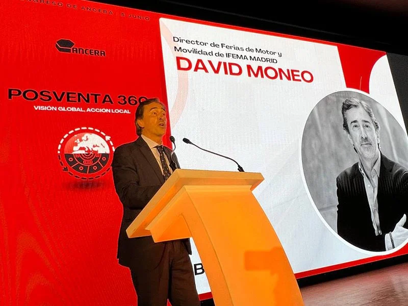 Un hombre habla en un podio en el escenario con una pantalla que muestra el nombre y la foto de David Moneo detrás de él.