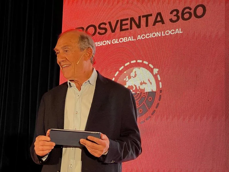 Hombre sonriente con traje sosteniendo una tableta, de pie frente a una pantalla de presentación roja "POSVENTA 360".