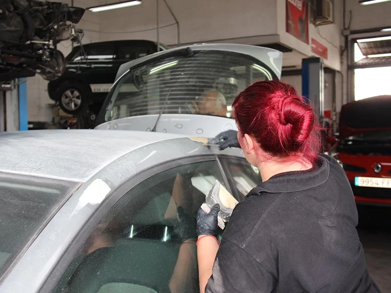 Una persona pelirroja lija el techo de un coche en un taller de reparación de automóviles, trabajando duro entre los vehículos mientras demuestran cómo podemos crecer juntos mediante el trabajo en equipo y la dedicación.