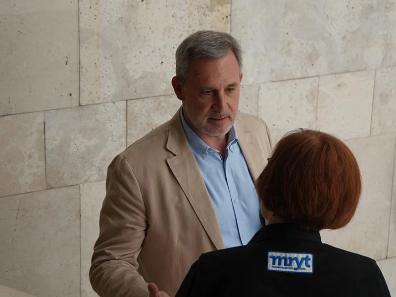 Un hombre con chaqueta beige habla con una mujer con chaqueta negra con el logotipo "mxyt", de pie contra un muro de piedra, mientras hablan de su viaje de pasos firmes.